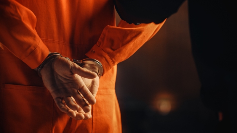 Person in an orange jumpsuit in handcuffs walking down a hallway
