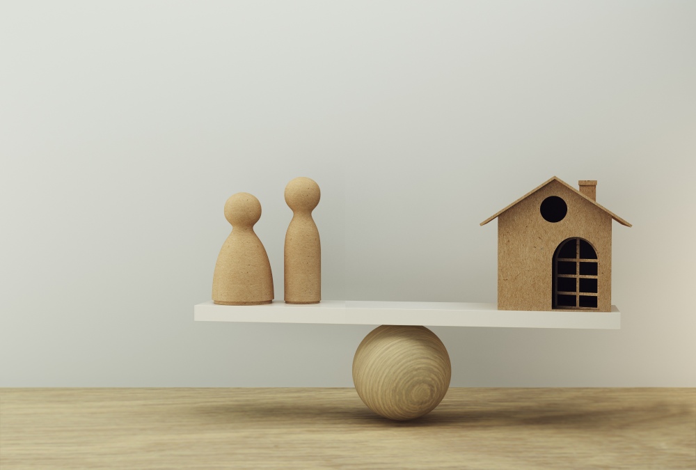Wooden home balances on seesaw with two wooden people on other end