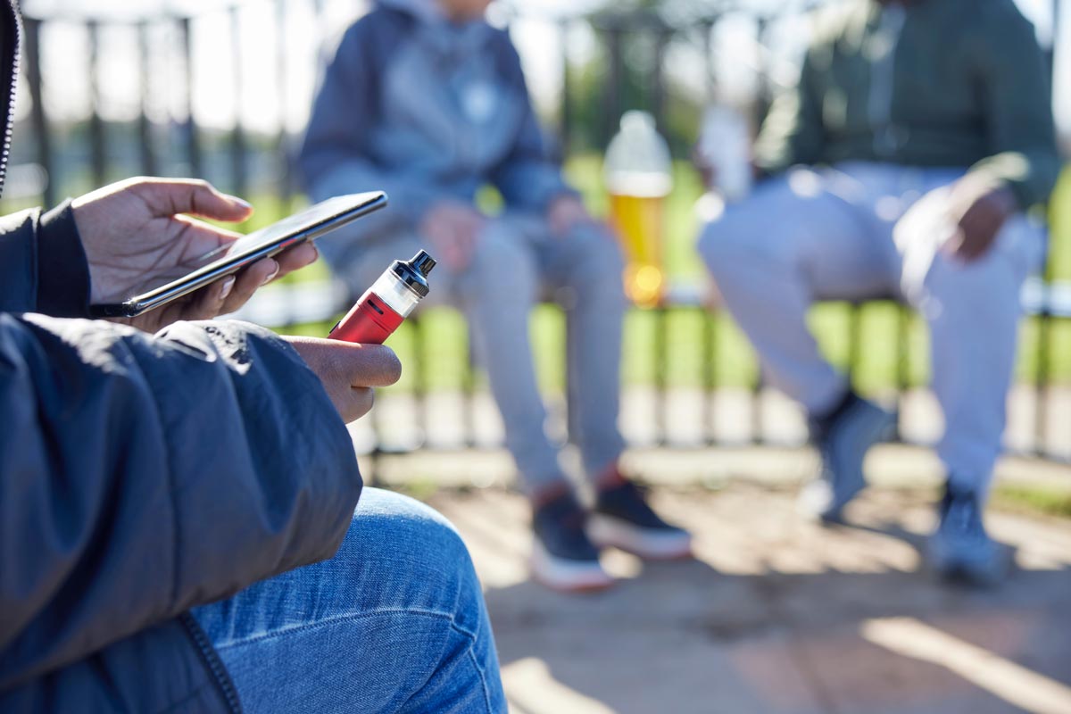 Young person in blue jeans holding mobile phone in left hand and vape device in right hand