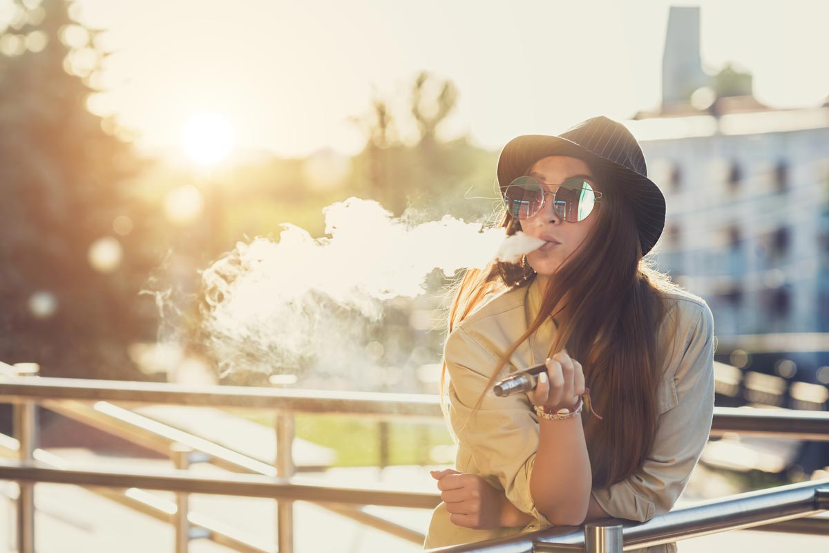Young woman with sunglasses and black hat vaping on a sunny day