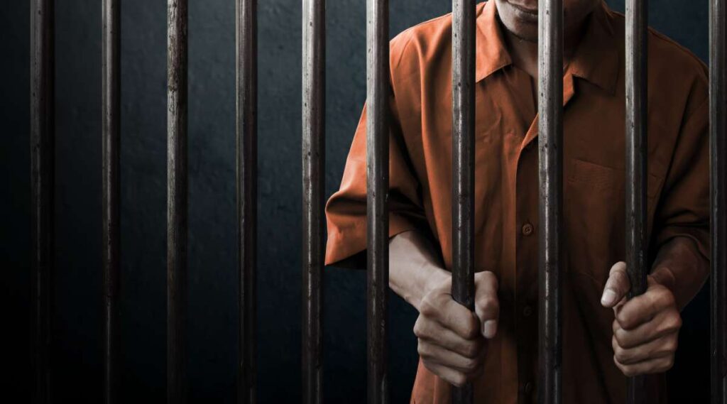 A man in an orange jumpsuit grips the bars of a poorly maintained jail cell in California