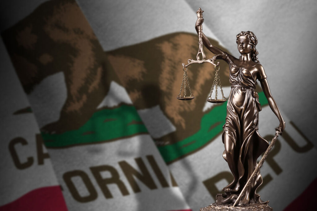 A statue of Lady Justice stands in front of a California state flag.