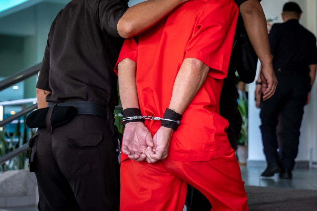 A man in a red prison jumpsuit in handcuffs, being escorted by police.